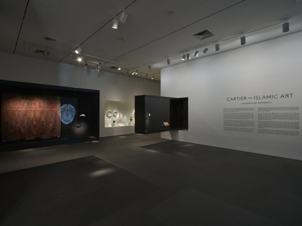 Exhibit room with dim lighting, displaying Cartier and Islamic Art with artifacts and text panels on the walls.