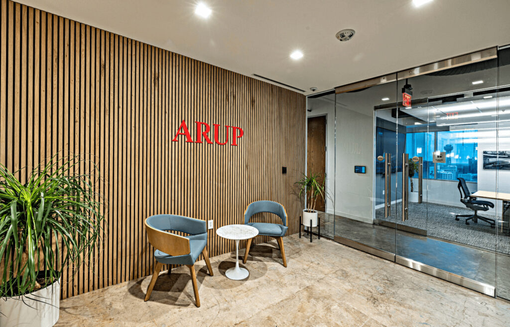 Office lobby with wooden slat wall, two blue chairs, and a small white table. Glass doors lead to a modern meeting room.