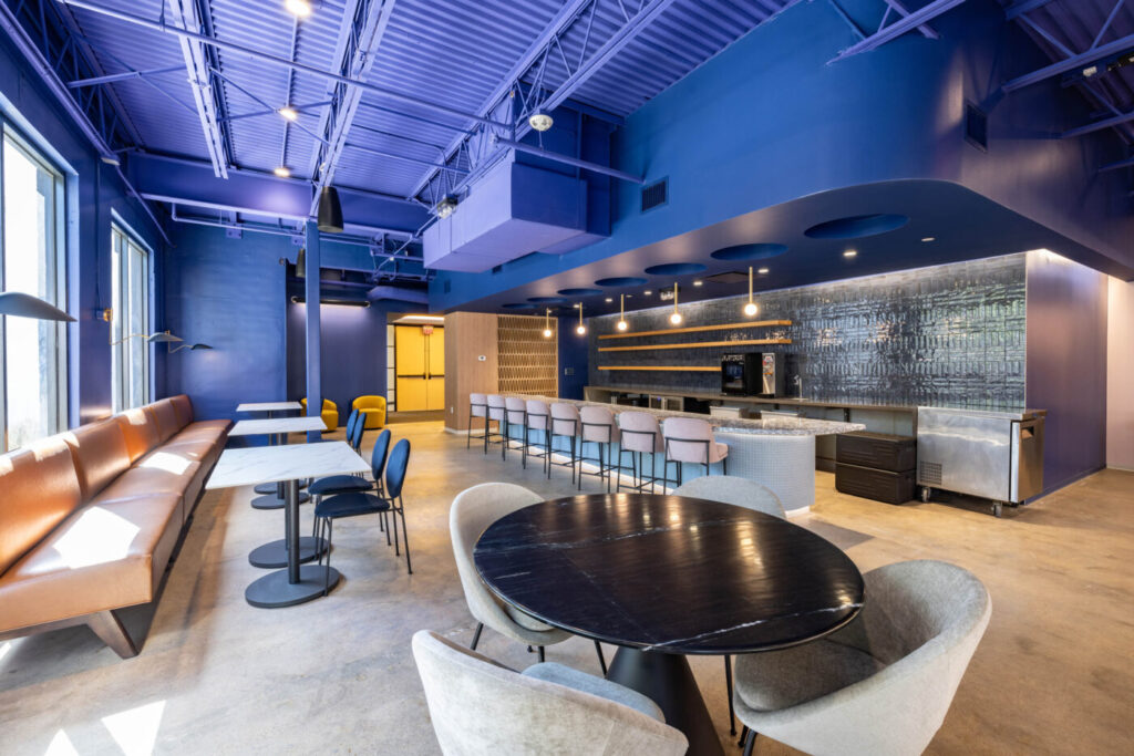 Modern lounge with blue walls, a social vibe, plush chairs, bench seating, and a long bar with stools. Bright natural light.