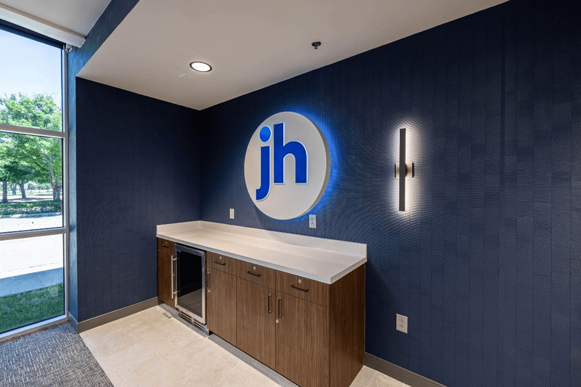 Office corner with a wooden cabinet, mini-fridge, and a blue logo on the wall. Natural light from a window illuminates the space.