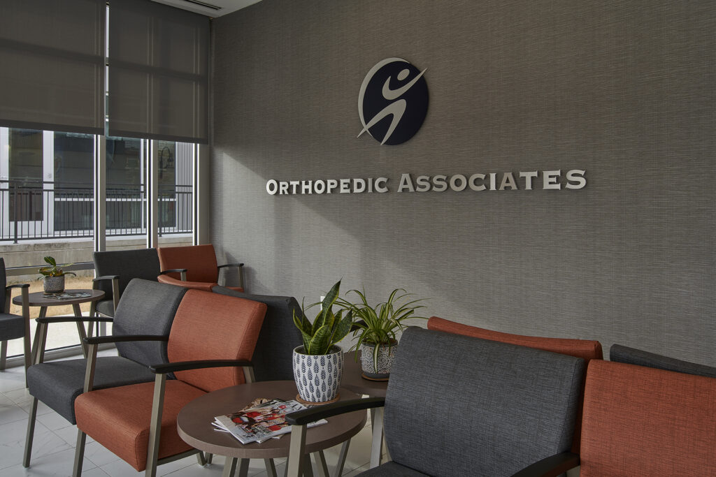 The waiting area has orange and gray chairs, small tables with plants, and a sign for FM Orthopedic Associates on the wall.
