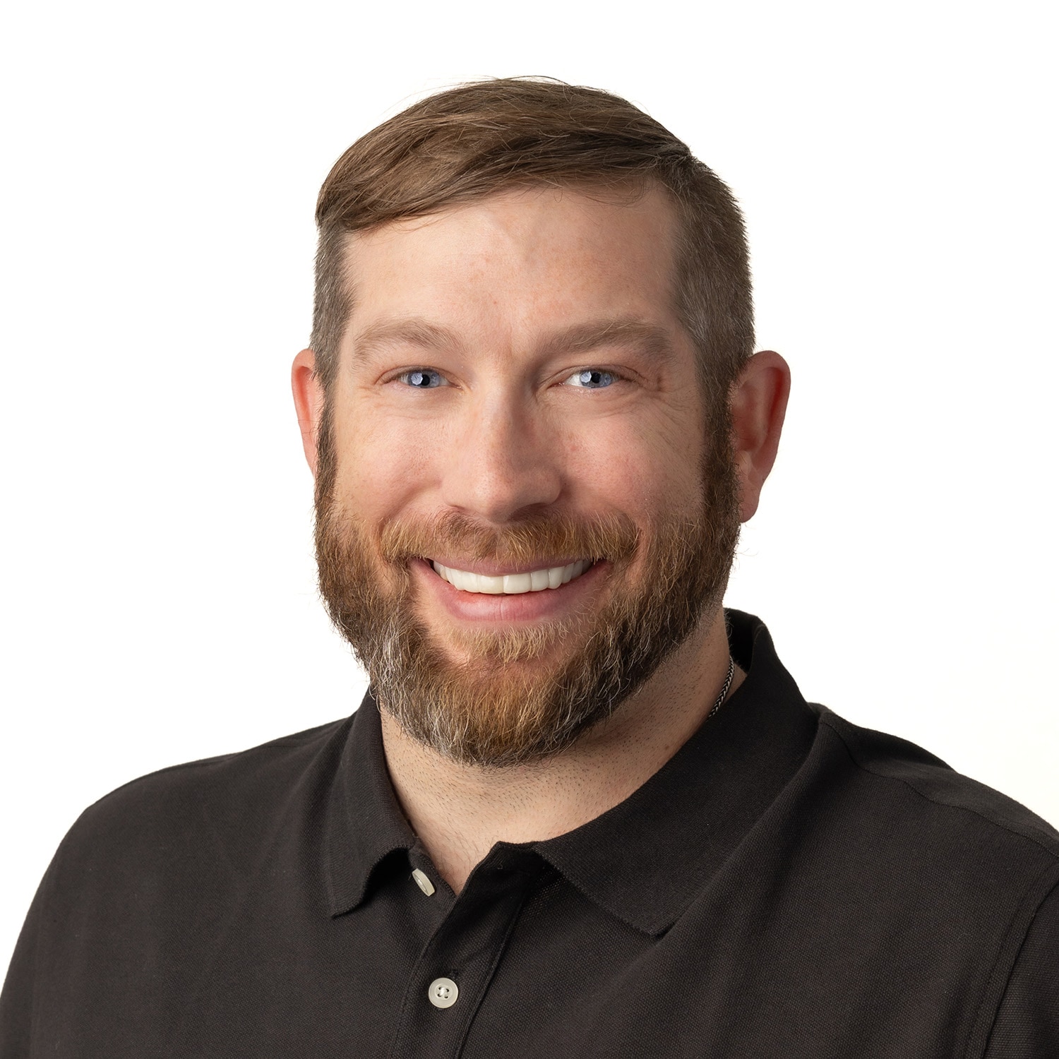 Smiling man with a beard and short hair, Daniel Behlar, in a black polo shirt against a white background.