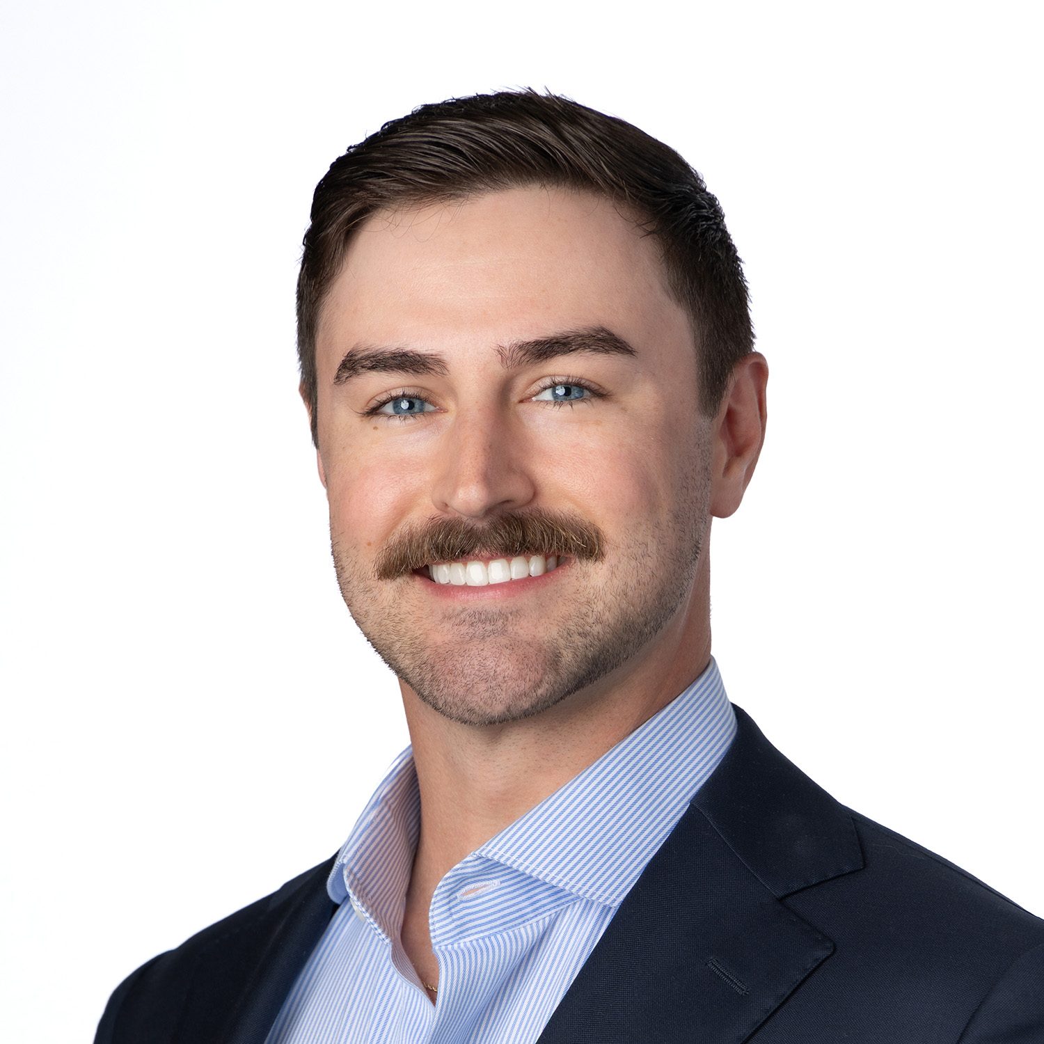 Man with brown hair, blue eyes, and a mustache, smiling in a suit jacket and striped shirt on a white background.